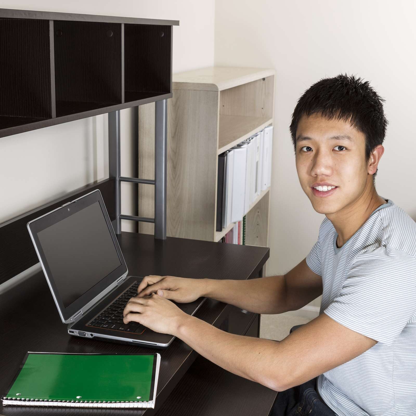 Young man studying at home office for college work with computer and notepad on desk